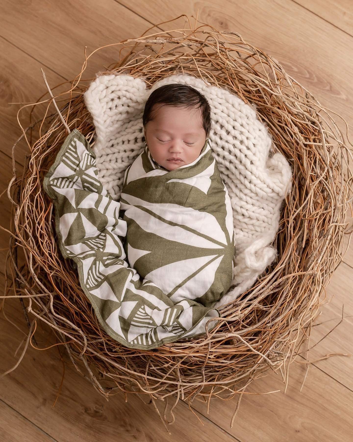 Baby wrapped in a sage green Polynesian inspired blanket inside of a straw nest