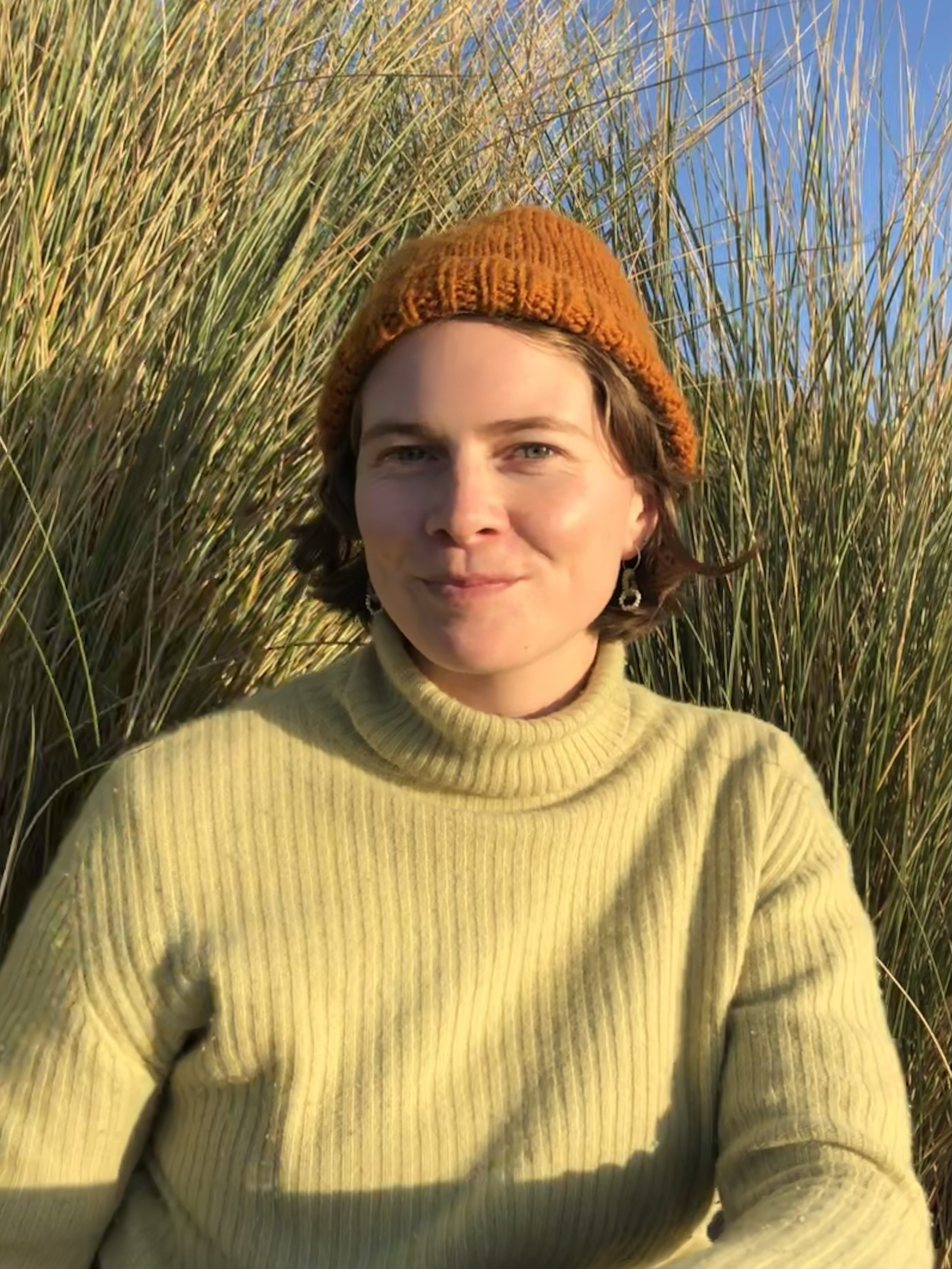 Avara Moody - Woman in green turtle neck and orange beanie in front of a green course bush